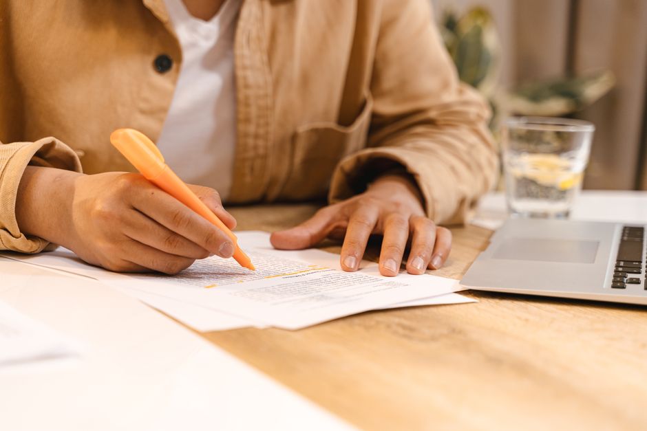 Osoba zaznaczająca pomarańczowym zakreślaczem tekst na kartce papieru przy laptopie.