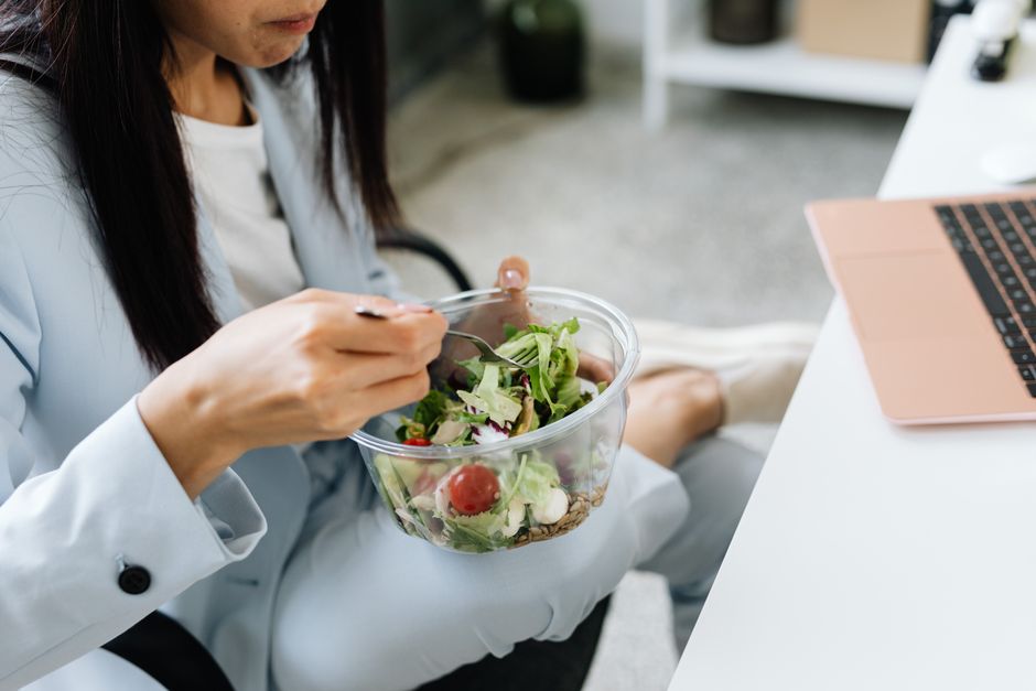 Kobieta w biurze je sałatkę z plastikowej miski przy laptopie