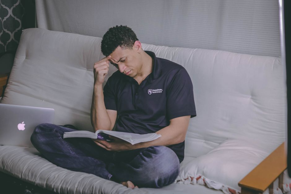 Mężczyzna siedzący na kanapie z otwartą książką i laptopem obok
