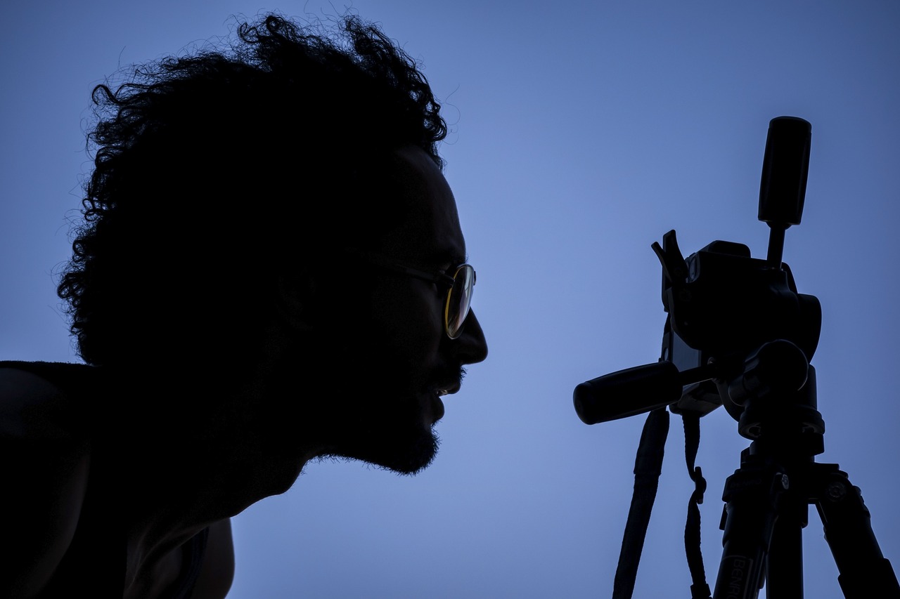Sylwetka fotografa w okularach patrzącego w aparat zamontowany na statywie